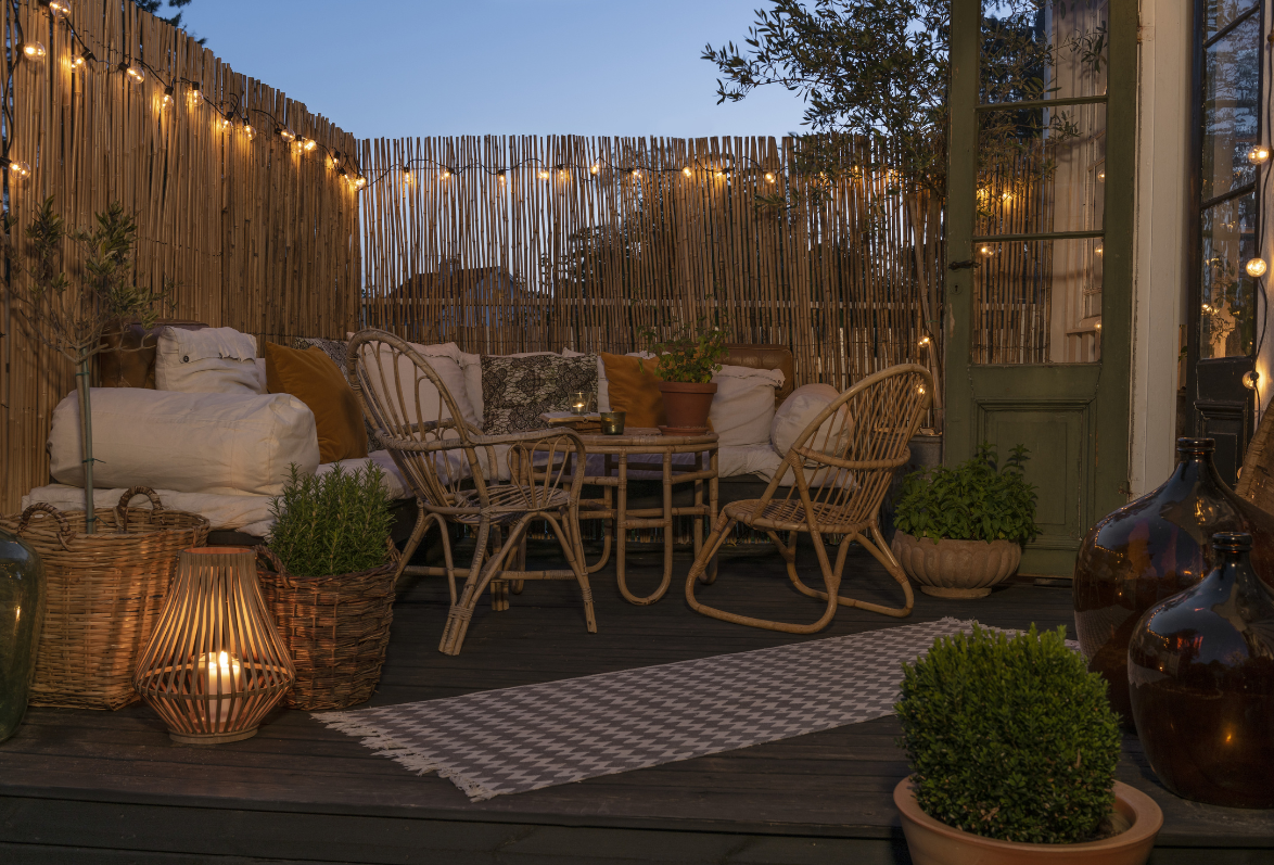 Festoon lights strung along a bamboo garden fence above a cosy patio seating area with lanterns and cushions at dusk