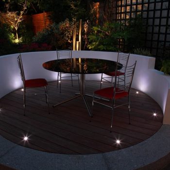 Curved rendered decking wall with small recessed ground lights around a round dining table and chairs with red cushions, lit at night