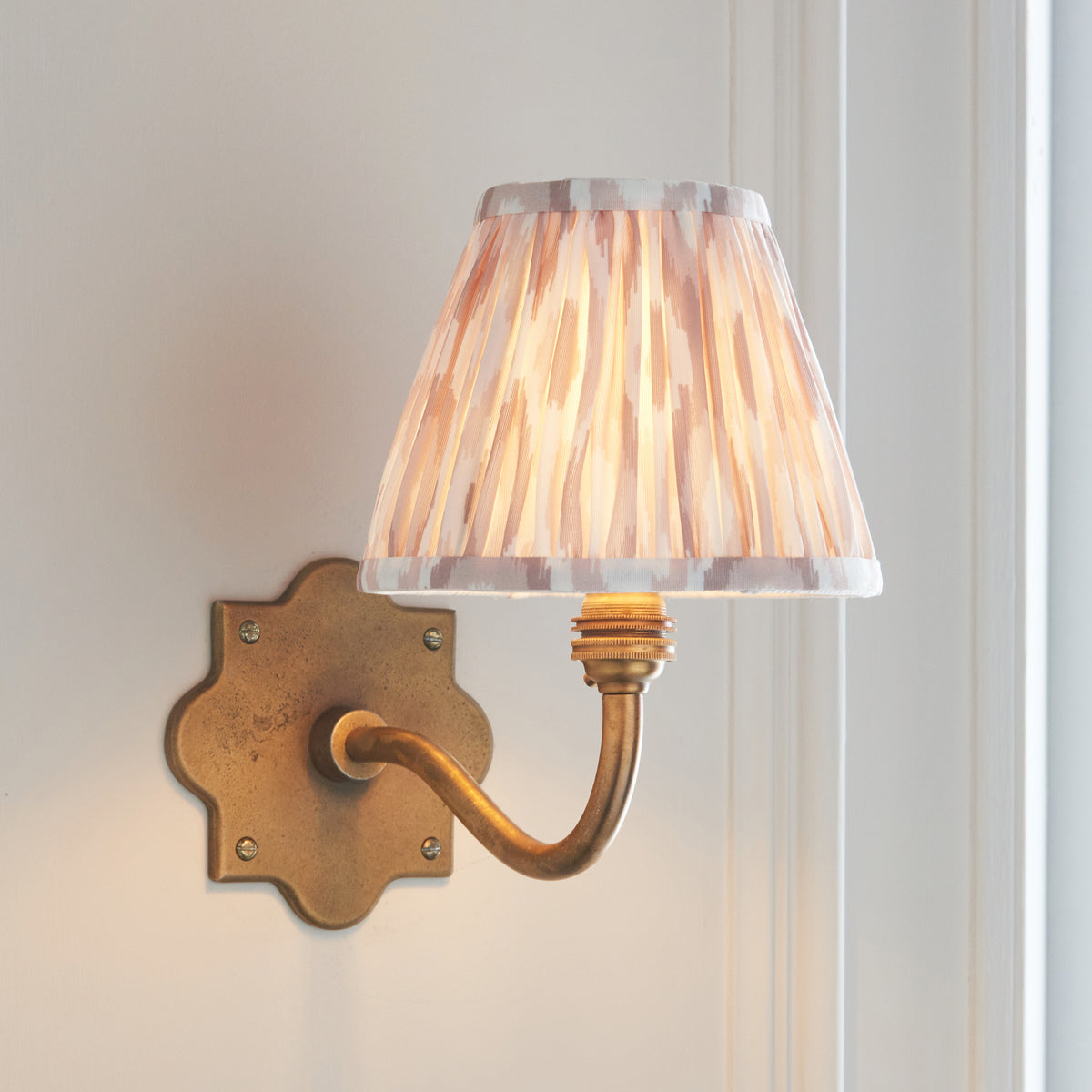 Endon Anvil Curve antique brass wall light with a neutral ikat shade, lit, on a pale wall beside a white door frame