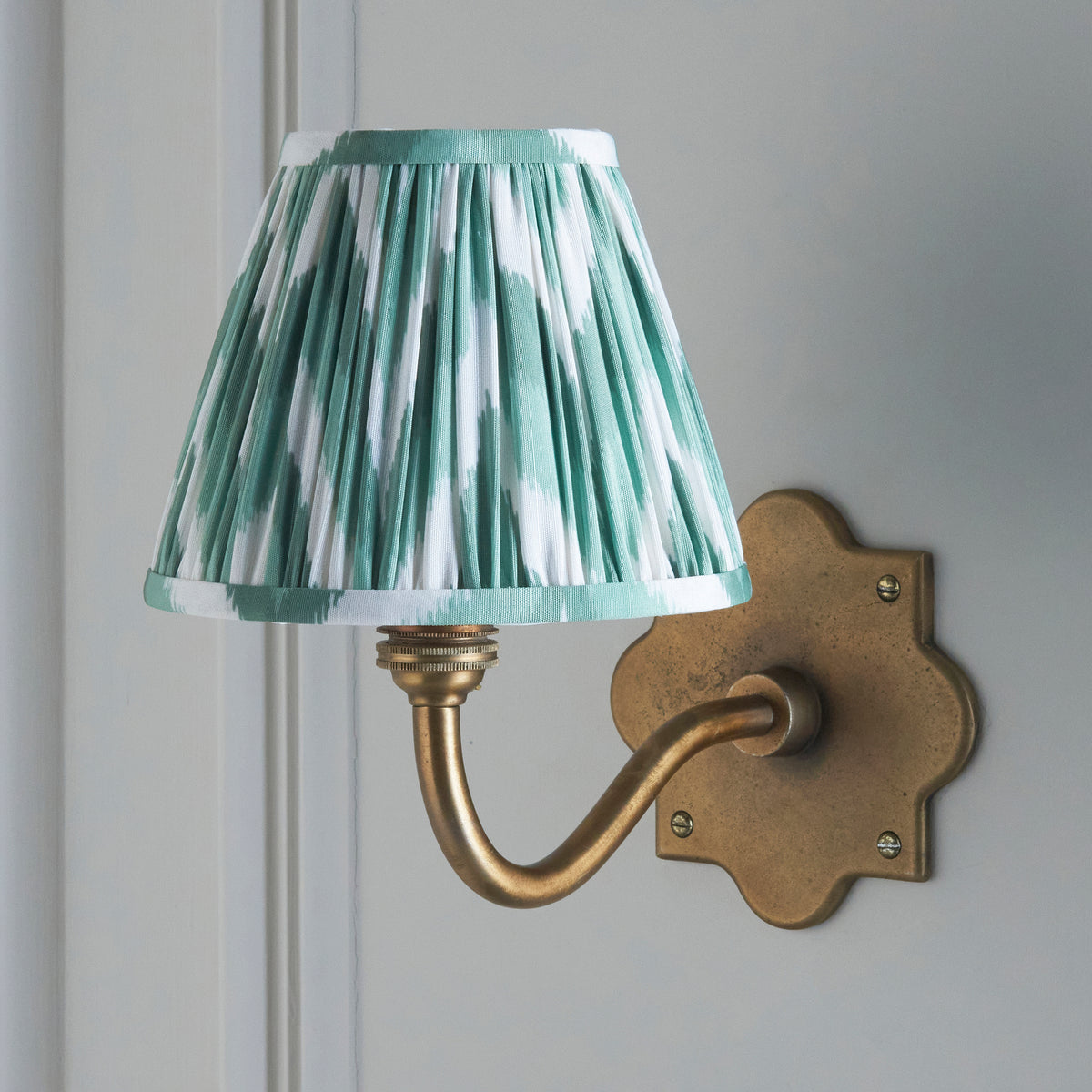 Endon Anvil Curve wall light unlit on a pale grey panelled wall, crisp green and white zigzag pleated shade over an antique brass arm