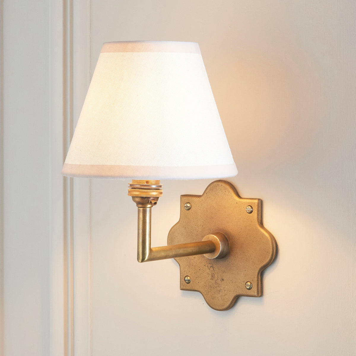 Endon Anvil Angle antique brass wall light with vintage white shade lit on a cream panelled wall, arm angled towards the viewer