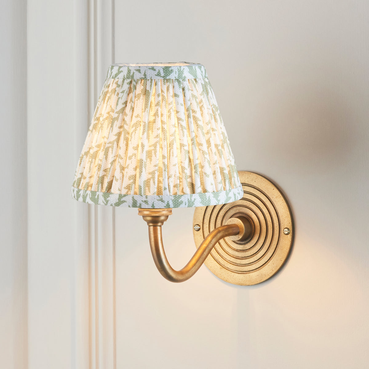 Endon Ripple Curve antique brass wall light lit on a cream panelled wall, green leaf pleated shade glowing warmly, ringed backplate visible