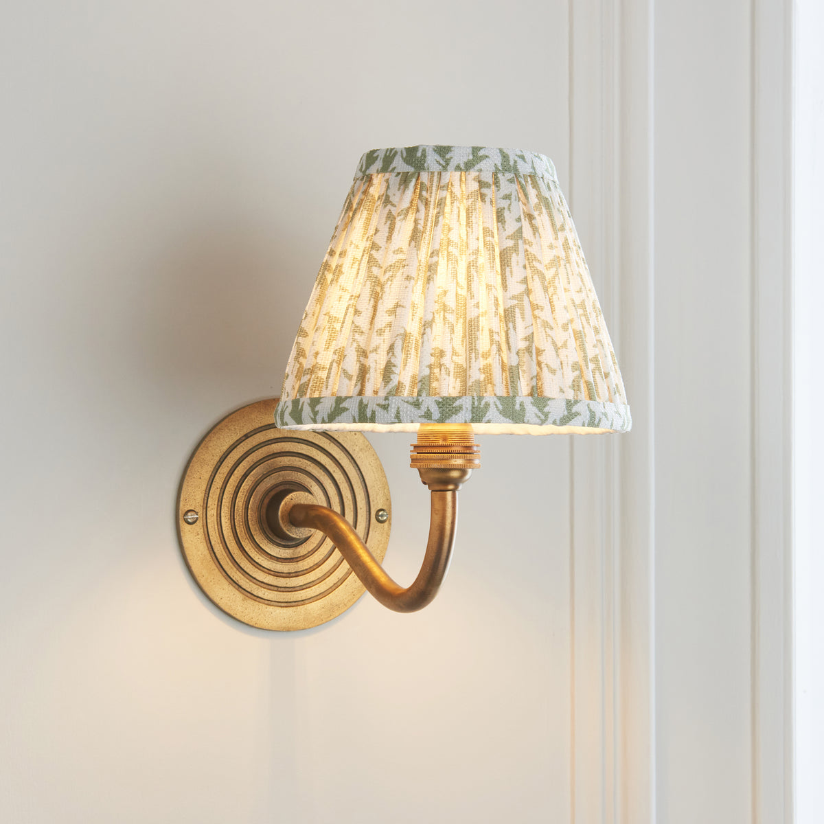 Endon Ripple Curve wall light lit on a white panelled wall, curved antique brass arm sweeping from the ringed backplate up to the leaf print shade