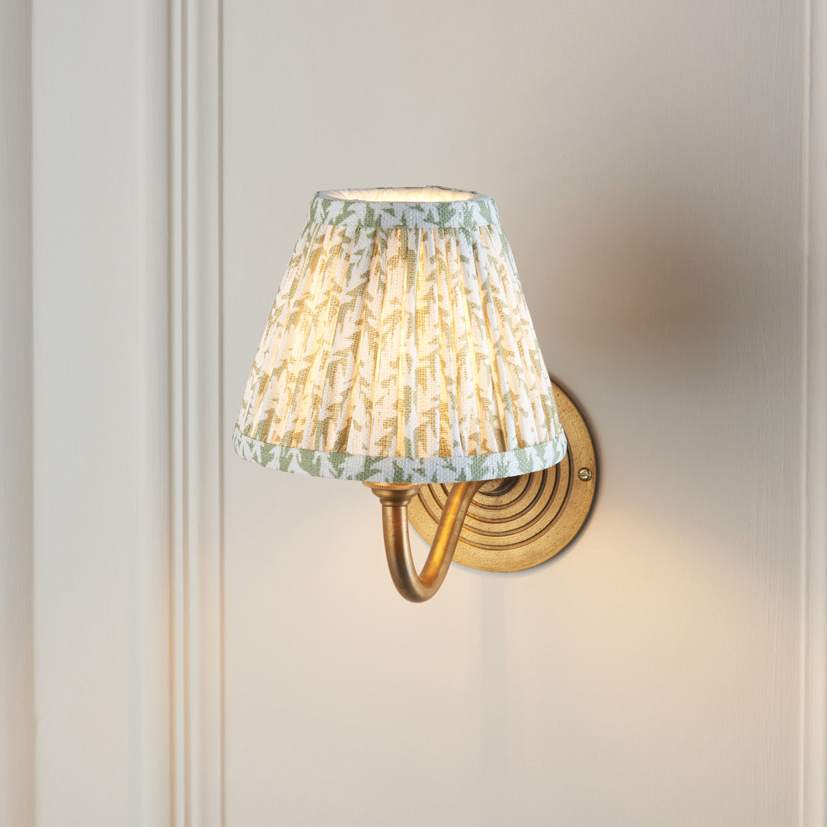 Endon Ripple Curve wall light switched on against a cream panelled wall, soft focus shot with the green leaf shade glowing and brass arm below