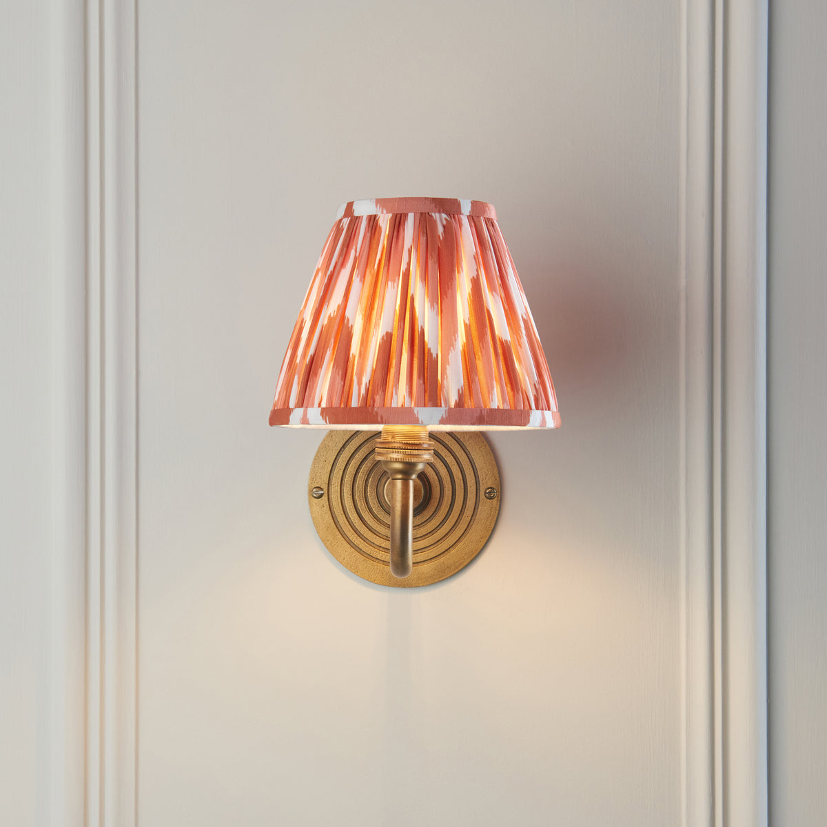 Endon Ripple Curve wall light switched on against cream wall panelling, orange and white zigzag shade sitting centrally above the antique brass backplate