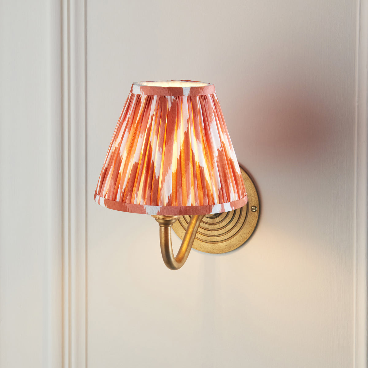Endon Ripple Curve wall light lit on a cream panelled wall with the open top of the apricot orange zigzag pleated shade showing