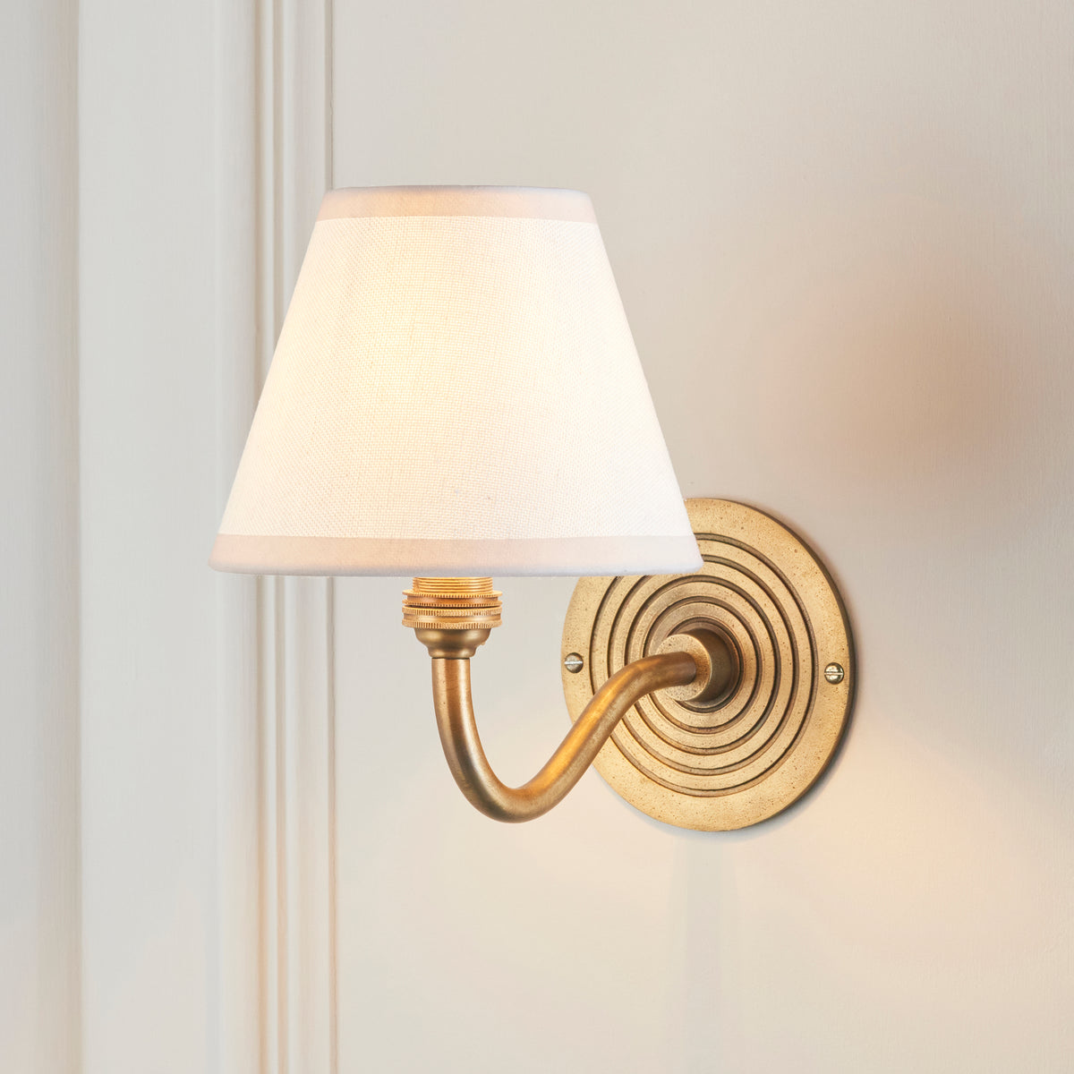 Endon Ripple Curve antique brass wall light with vintage white shade, lit, mounted on a cream panelled wall