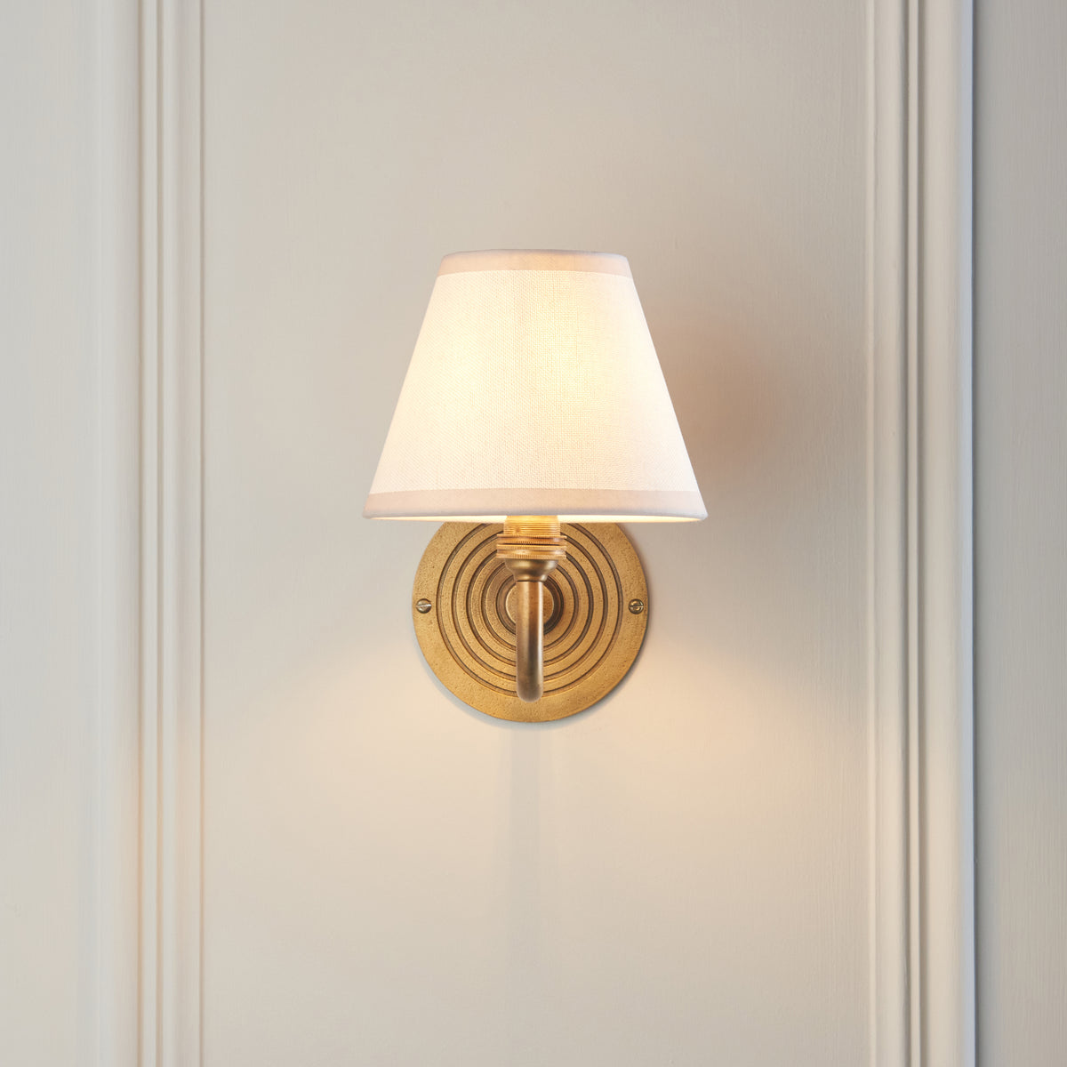 Endon Ripple Curve wall light lit on a cream panelled wall, showing the ringed antique brass backplate below the vintage white shade