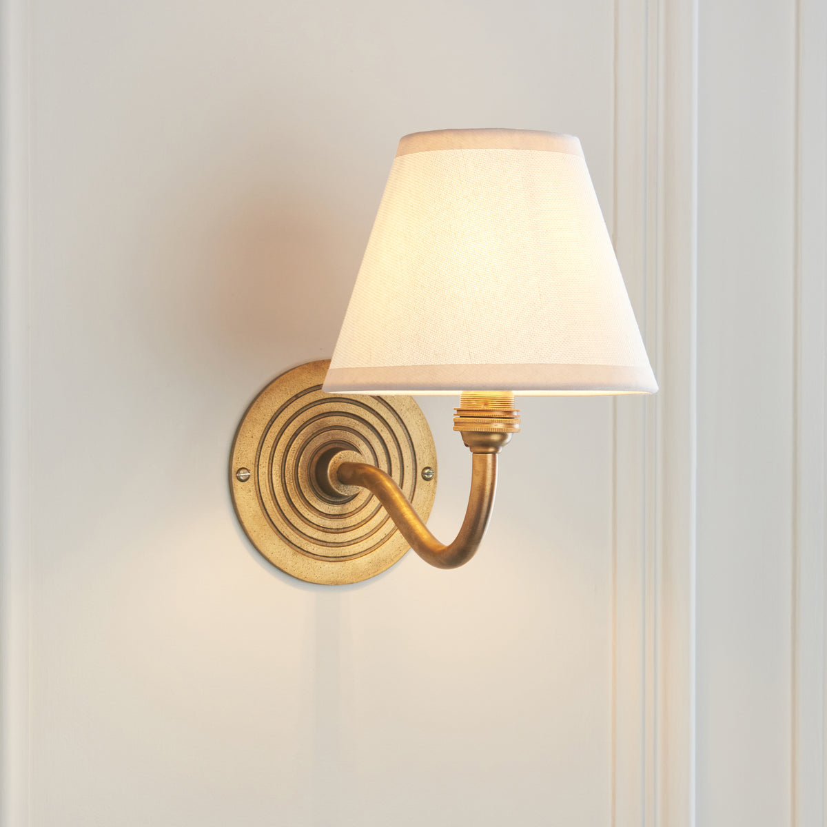 Endon Ripple Curve antique brass wall light lit on a cream panelled wall, curved arm and ringed backplate beside the white shade