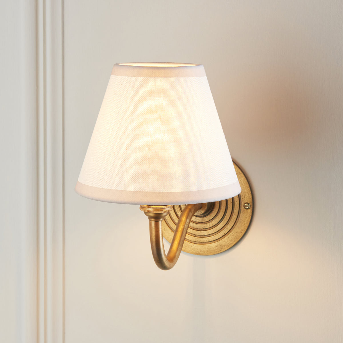 Endon Ripple Curve wall light lit on a cream wall, vintage white shade glowing with warm light spreading around the brass backplate