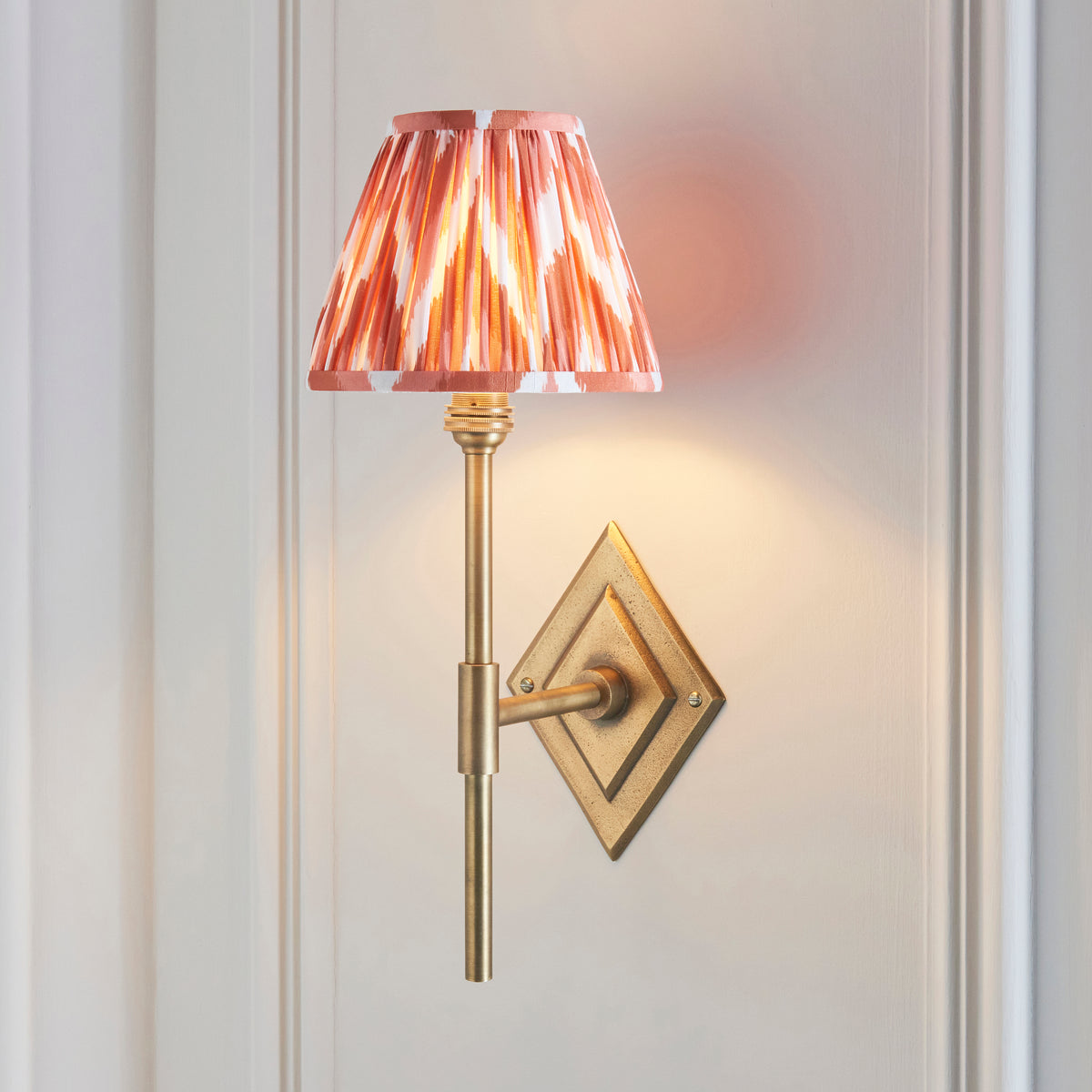 Endon Diamond Tall wall light lit on a pale grey panelled wall, antique brass diamond backplate and apricot orange zigzag pleated shade