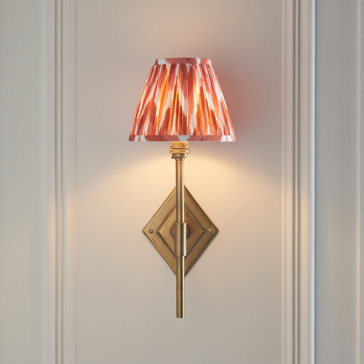 Endon Diamond Tall antique brass wall light lit on a pale panelled wall, stem rising in front of the diamond backplate, apricot orange zigzag shade