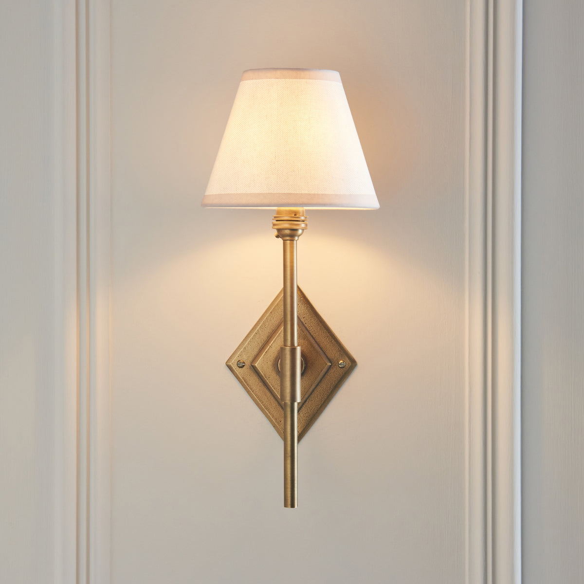 Lit Endon Diamond Tall wall light on cream wall panelling, stepped diamond backplate in antique brass and vintage white fabric shade