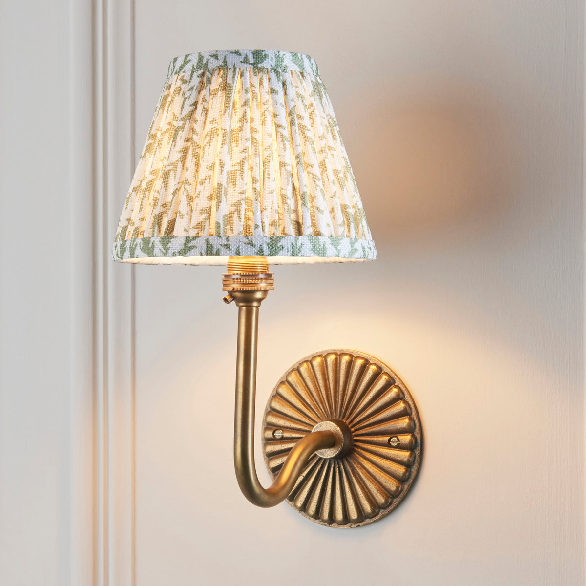 Endon Dahlia Swan antique brass wall light with green leaf pleated shade, lit, casting a warm glow on a cream panelled wall