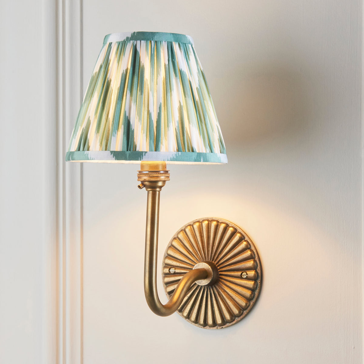 Endon Dahlia Swan antique brass wall light lit on a cream panelled wall, green zigzag shade above the ribbed backplate