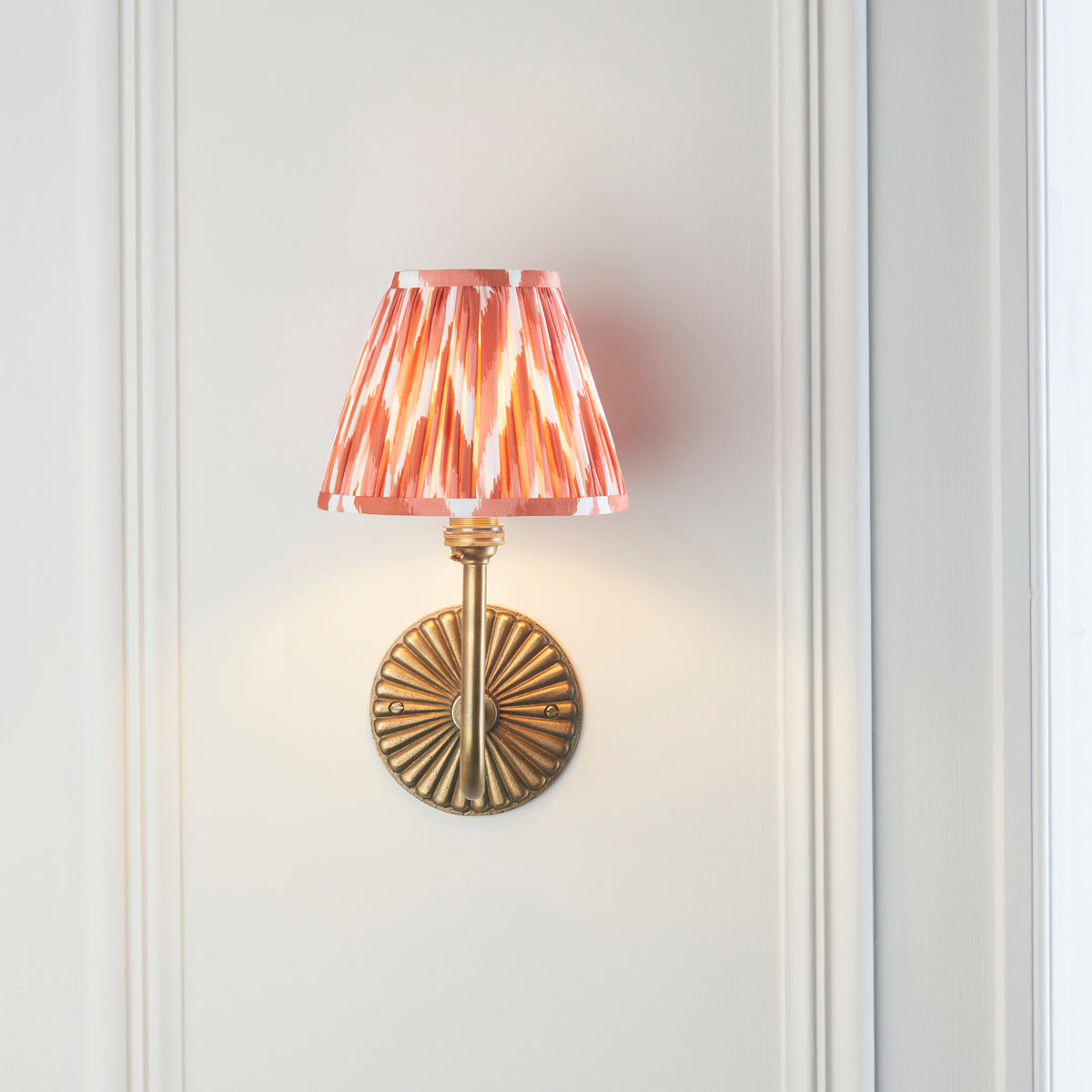Endon Dahlia Swan wall light lit on a pale panelled wall seen from a distance, small apricot orange shade over an antique brass backplate