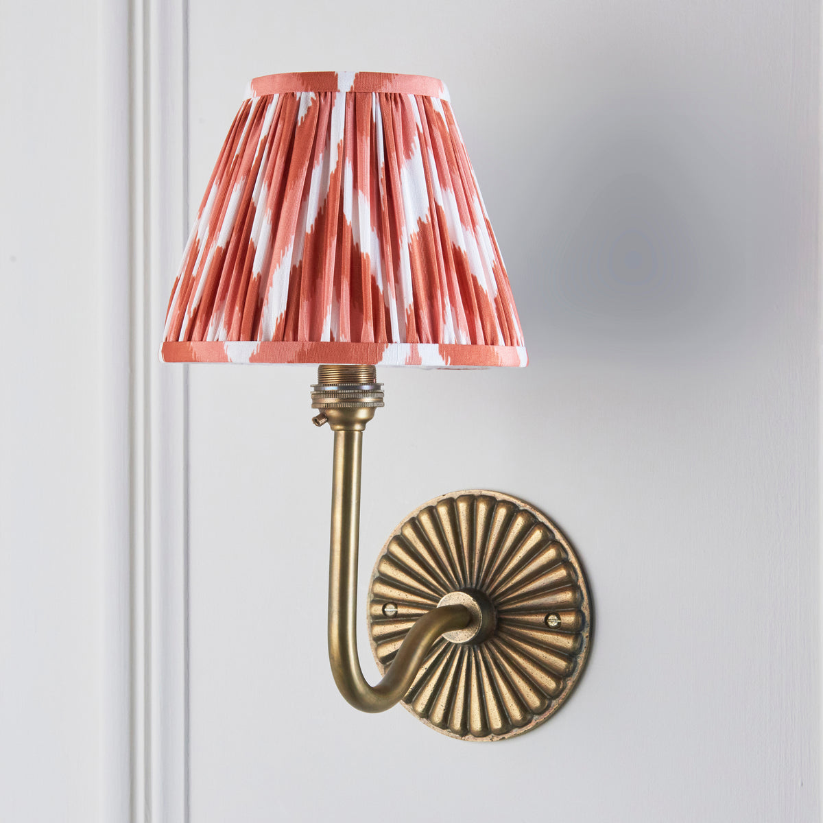 Endon Dahlia Swan wall light unlit on a pale grey panelled wall, apricot orange zigzag pleated shade and antique brass sunburst backplate