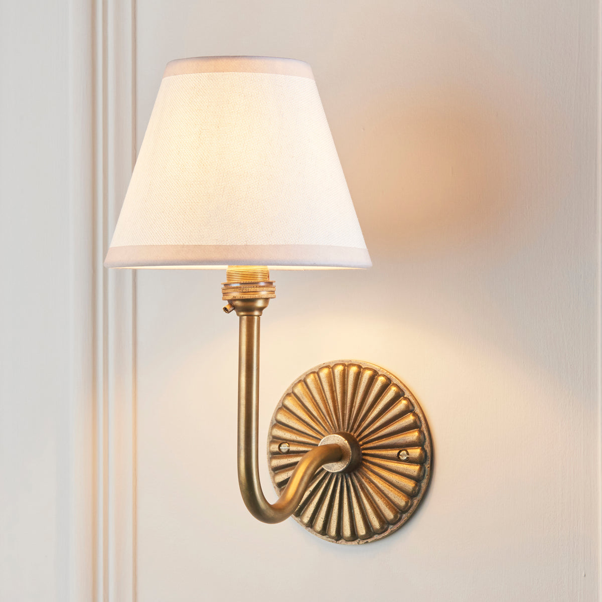 Endon Dahlia Swan antique brass wall light lit on a cream panelled wall, vintage white shade glowing softly