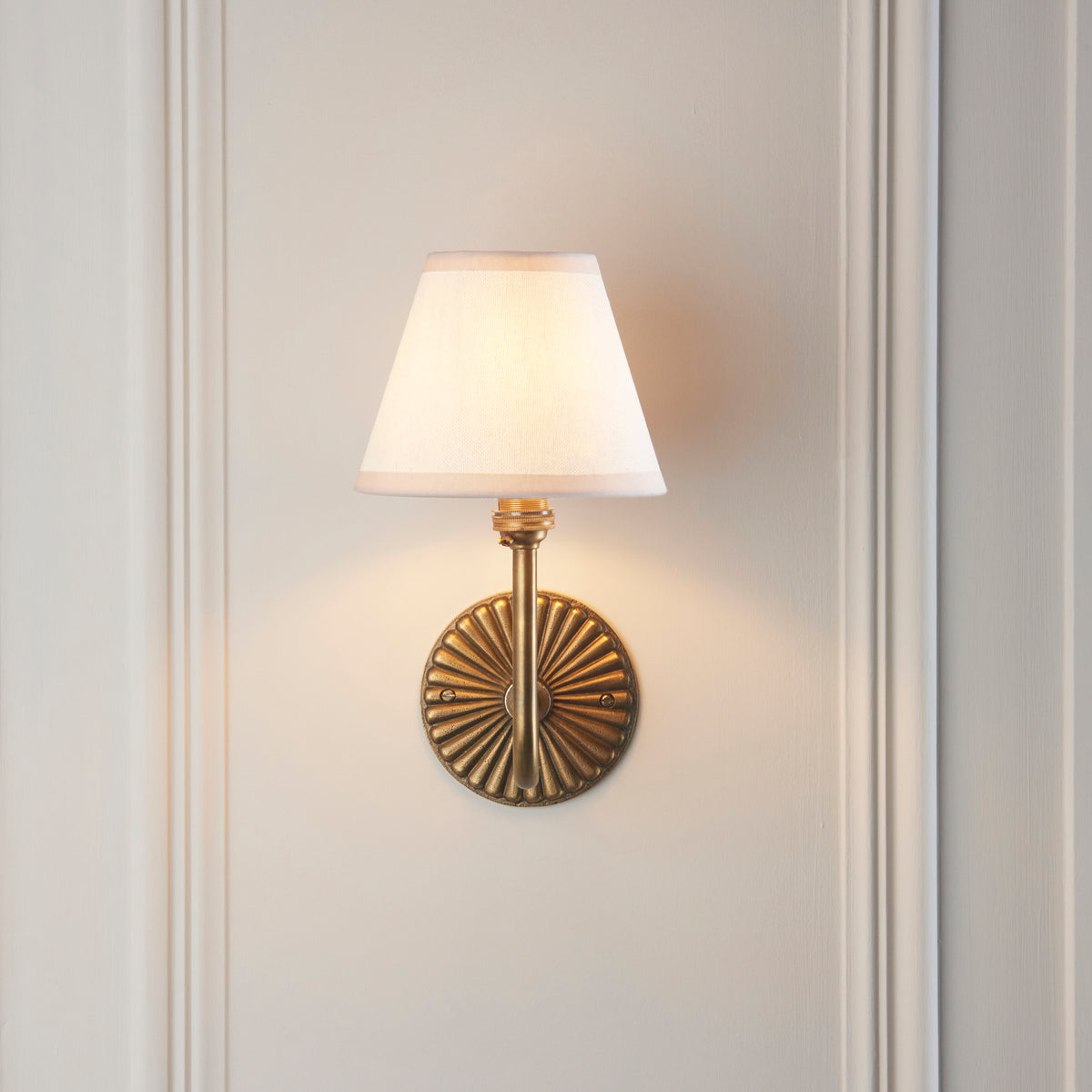 Endon Dahlia Swan wall light lit on a cream panelled wall, the brass arm rising in front of the fluted backplate