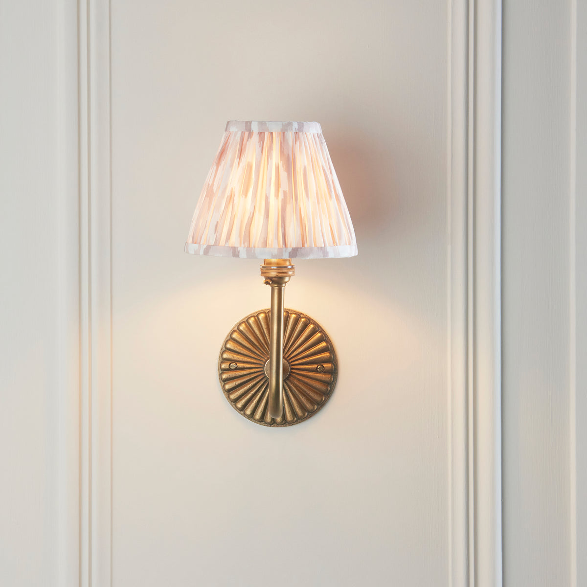 Endon Dahlia Swan wall light lit on a cream panelled wall, ribbed antique brass backplate below the pleated neutral ikat shade