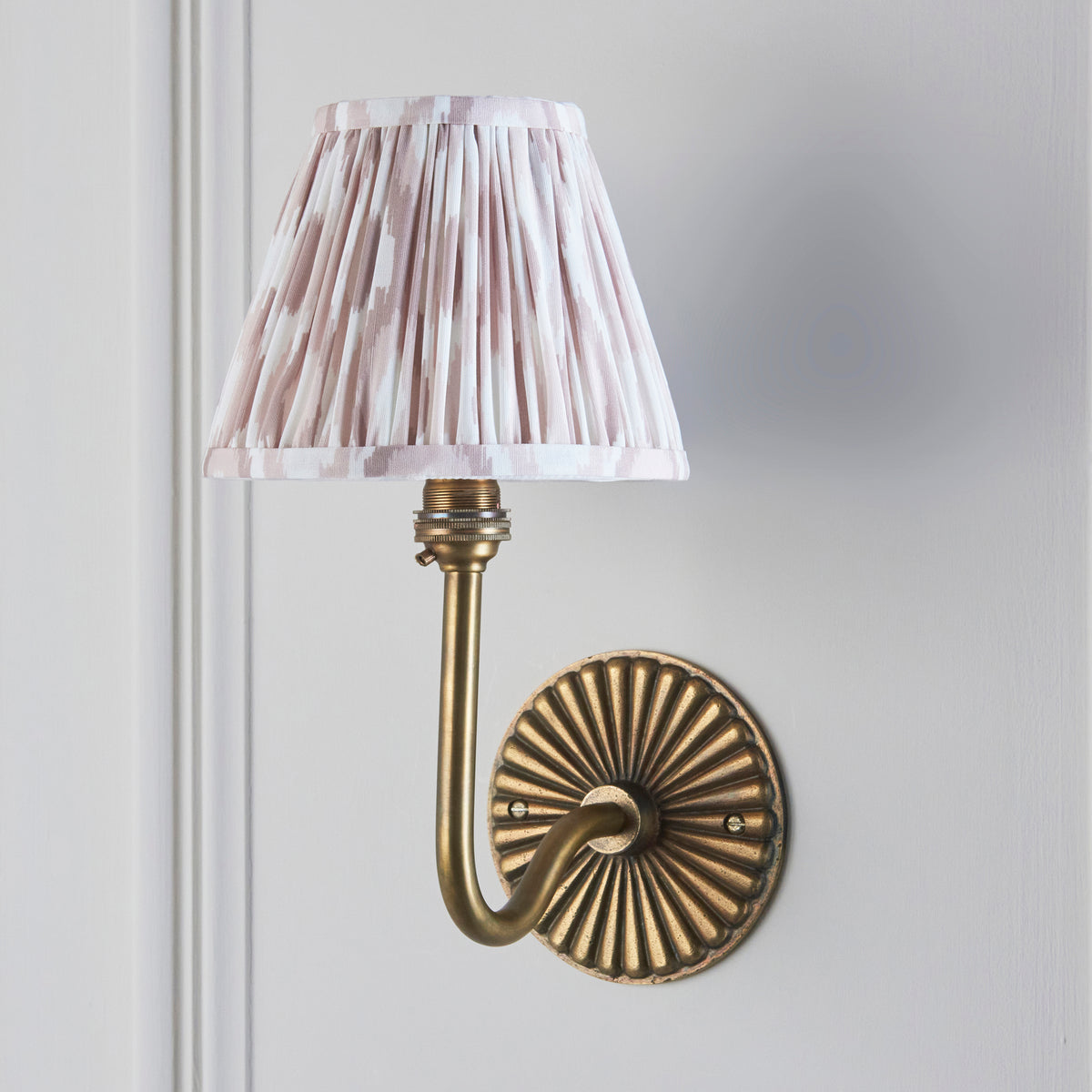 Endon Dahlia Swan wall light unlit on a pale grey panelled wall, antique brass bracket with pleated pink ikat fabric shade