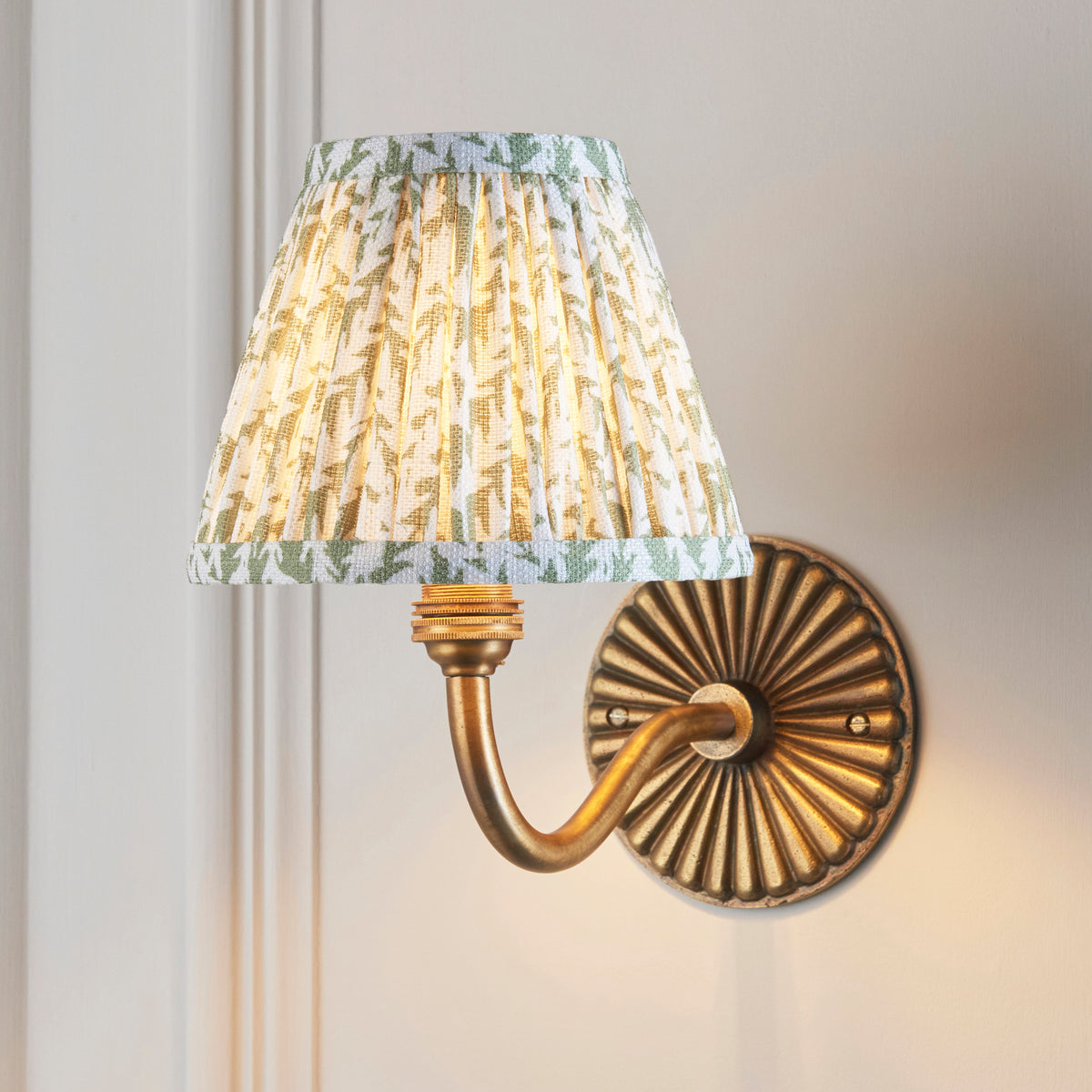 Endon Dahlia Curve wall light lit on a cream panelled wall, green leaf pleated shade glowing above the fluted antique brass backplate