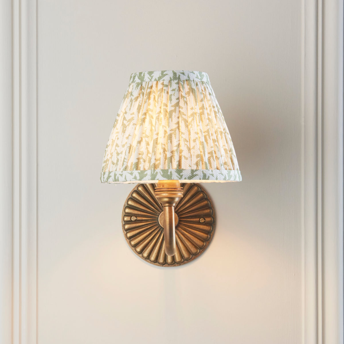 Endon Dahlia Curve antique brass wall light lit on a cream panelled wall with the fluted backplate directly beneath the green leaf shade
