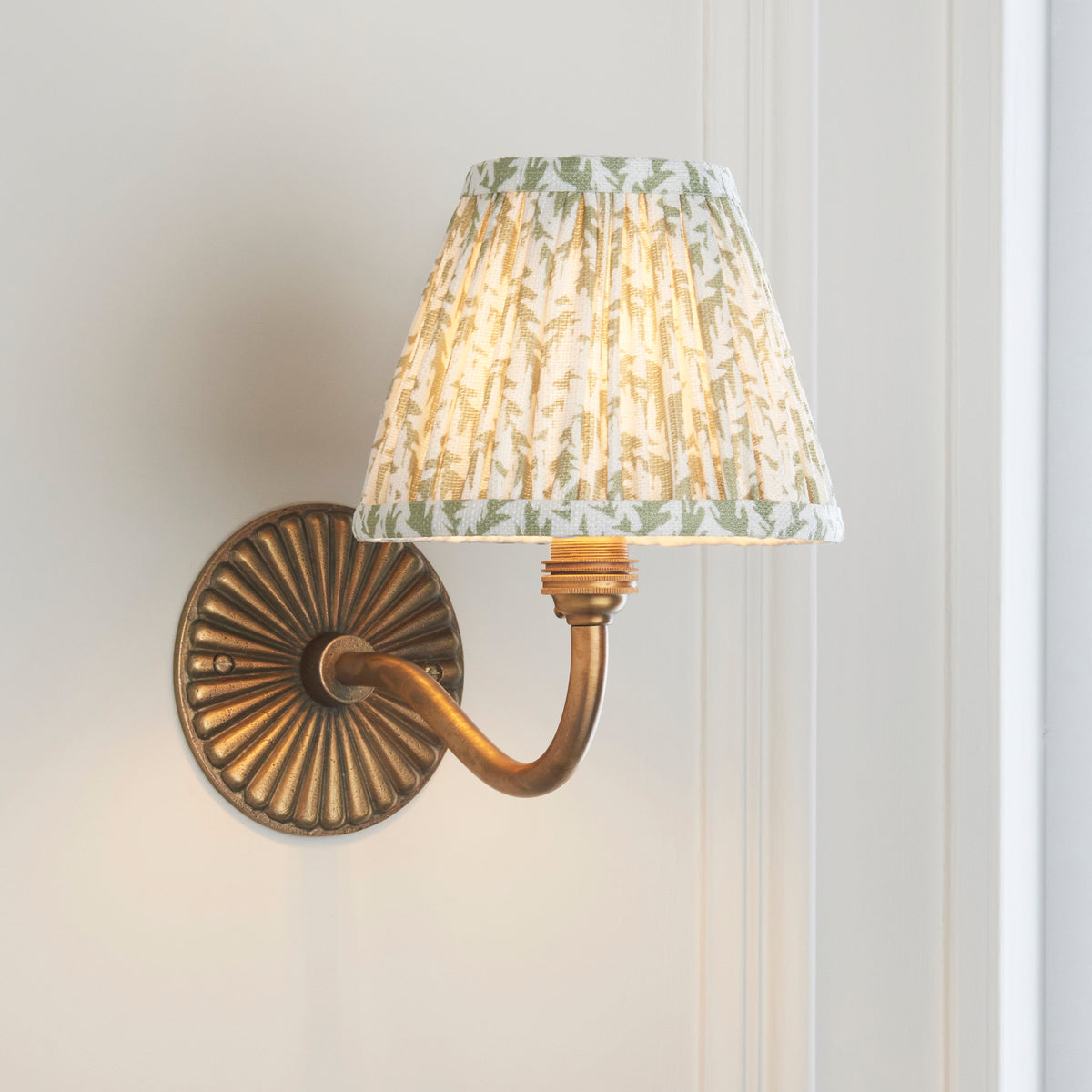 Endon Dahlia Curve wall light lit on a cream panelled wall beside a white door frame, brass arm sweeping up to the green leaf shade