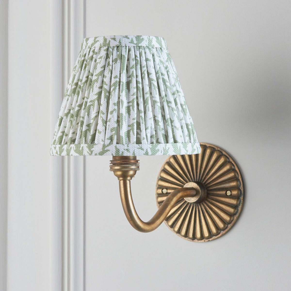 Endon Dahlia Curve antique brass wall light unlit on a cream panelled wall, green and off white leaf print pleated shade