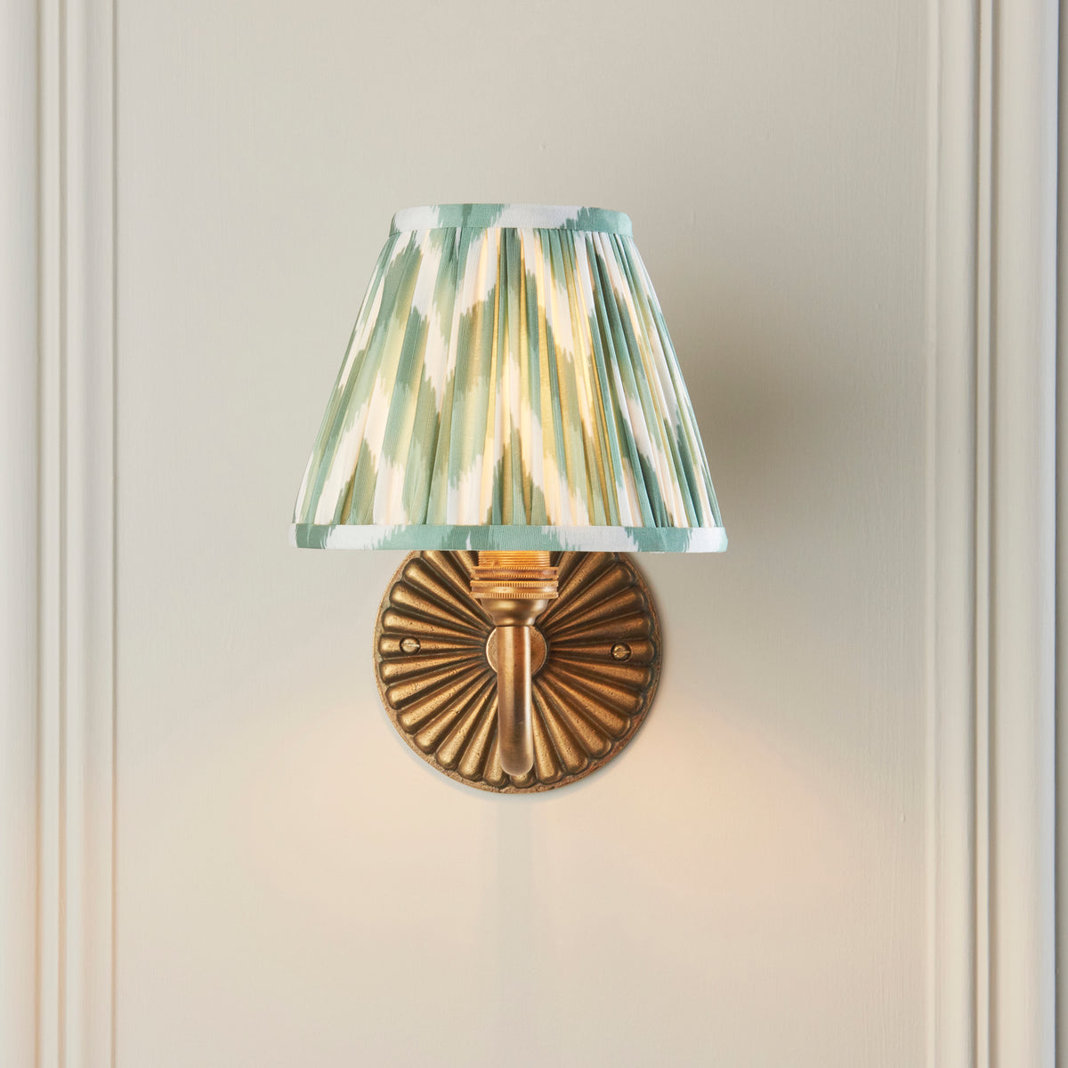 Endon Dahlia Curve wall light lit on a cream panelled wall, Cotswold green zigzag shade sitting above the fluted brass backplate