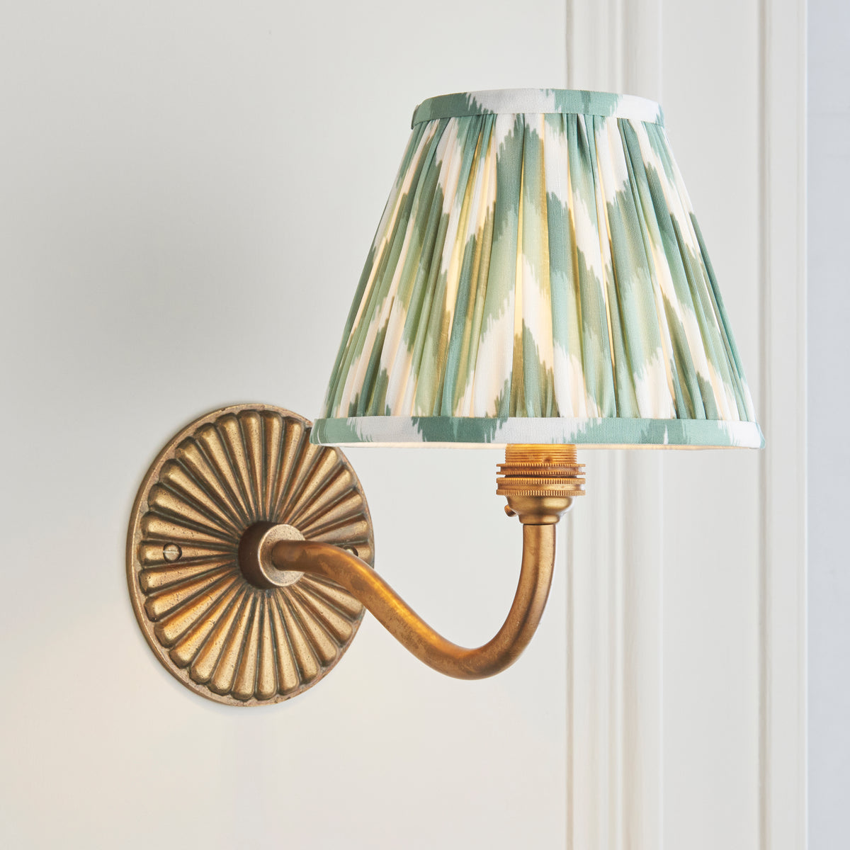 Endon Dahlia Curve wall light lit in a white panelled room, antique brass arm curving out beneath the green zigzag shade