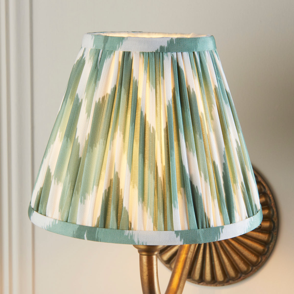Close up of the lit Cotswold green zigzag pleated shade on the Endon Dahlia Curve wall light, brass backplate behind