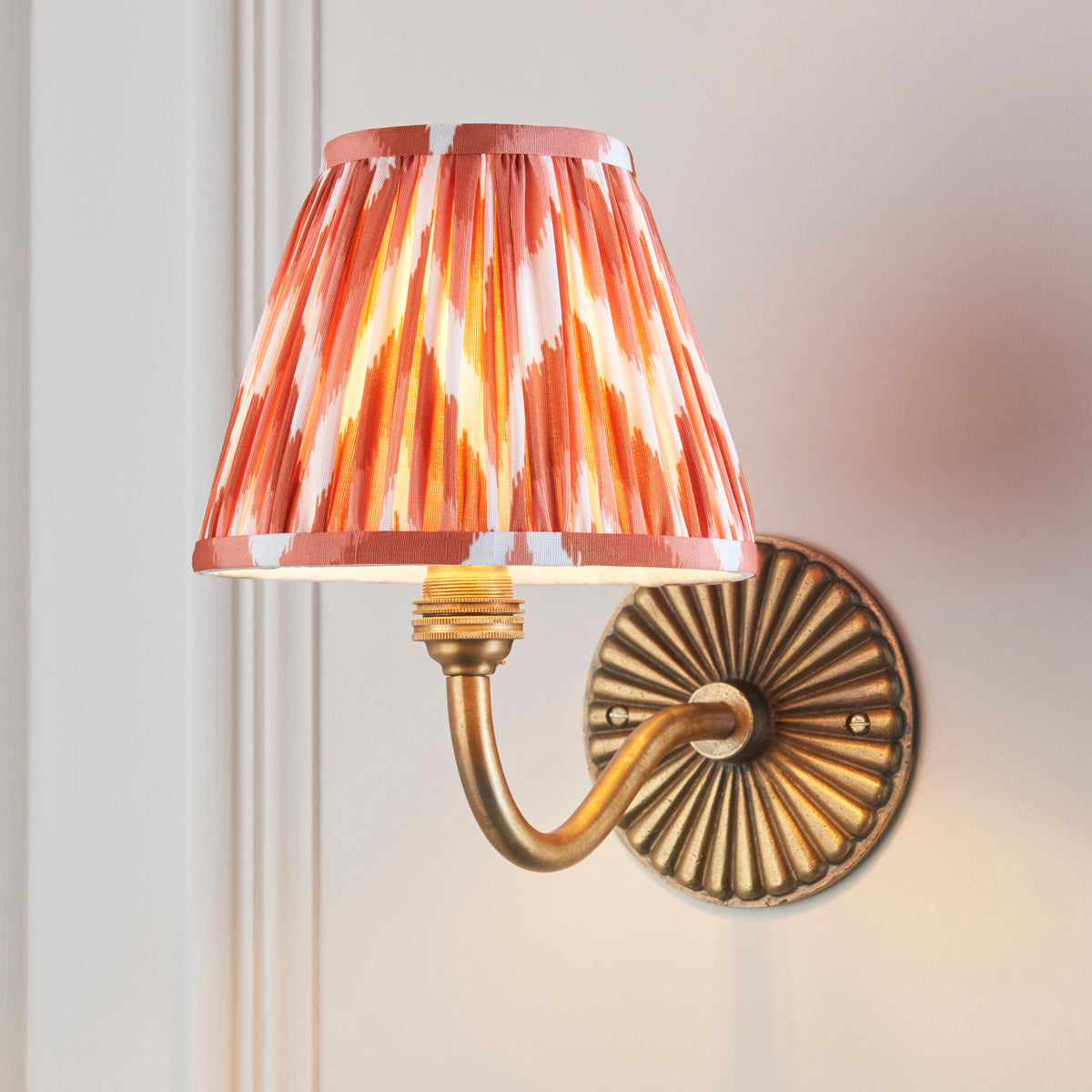 Endon Dahlia Curve antique brass wall light lit on a cream panelled wall, apricot orange zigzag shade glowing