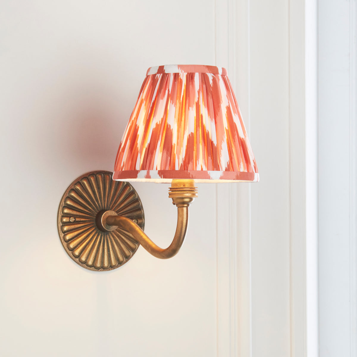Endon Dahlia Curve antique brass wall light lit on a cream panelled wall, curved arm and apricot orange zigzag shade with wall space around it