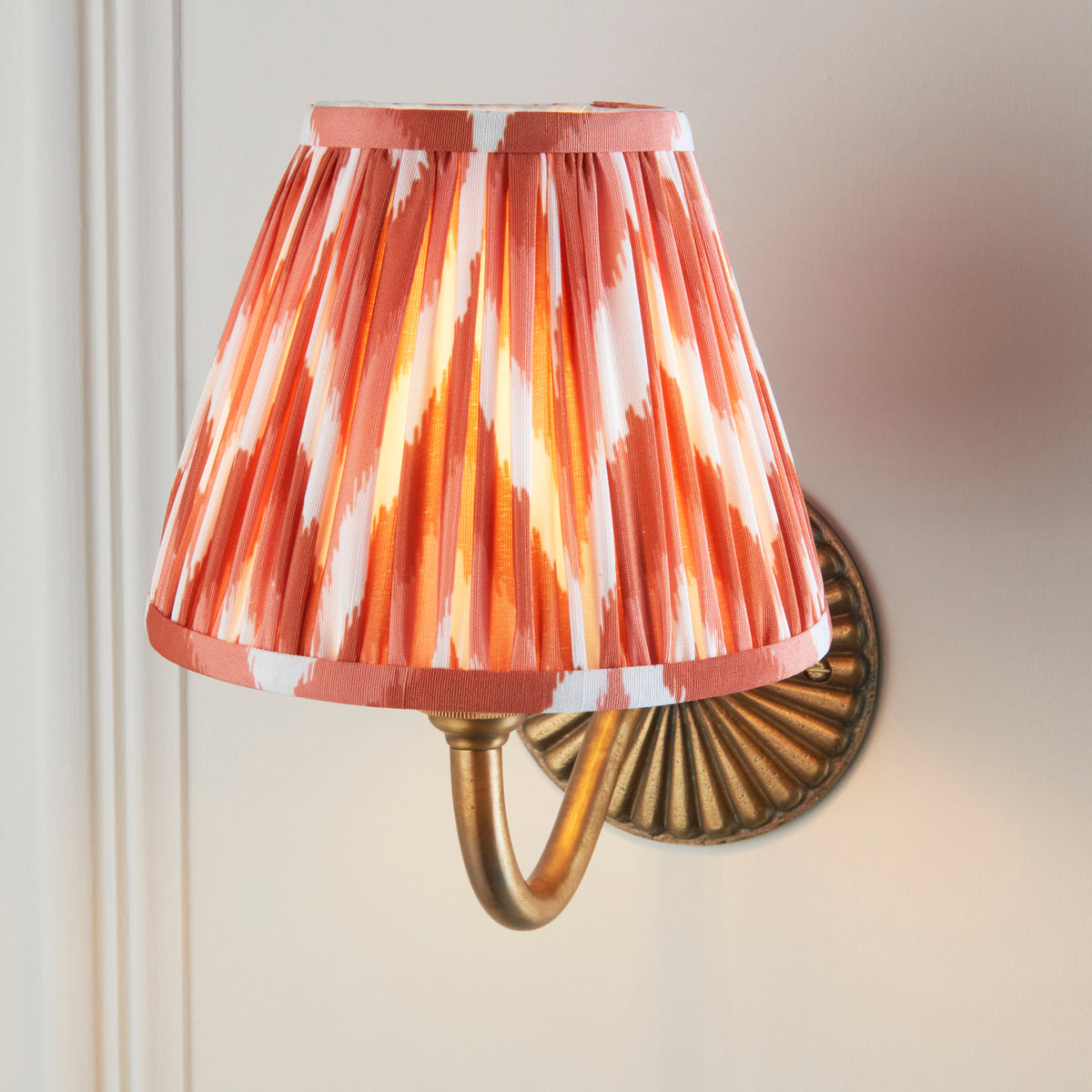 Close up of the Endon Dahlia Curve wall light lit, apricot orange zigzag pleated shade glowing in front of the antique brass backplate