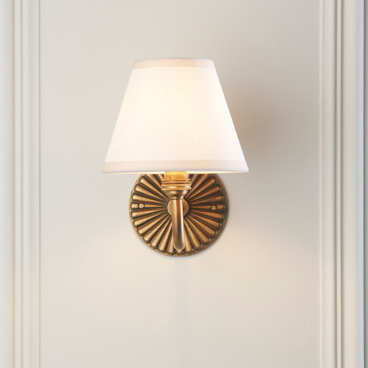 Endon Dahlia Curve wall light lit on a cream panelled wall, the round fluted antique brass backplate showing beneath the vintage white shade