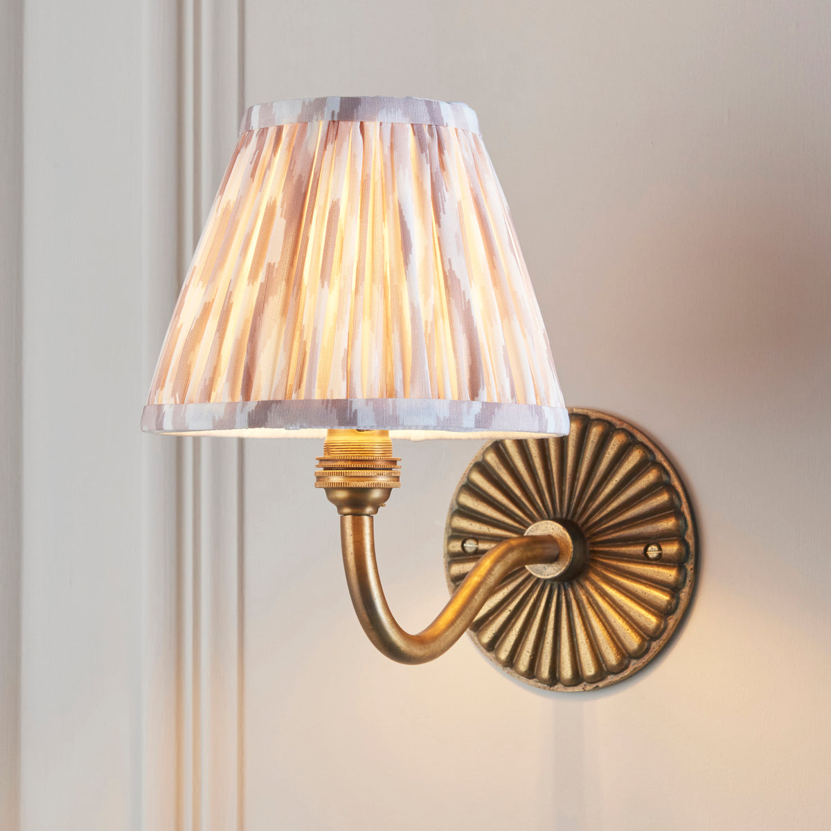 Endon Dahlia Curve antique brass wall light with ikat shade lit on a cream panelled wall, curved arm visible