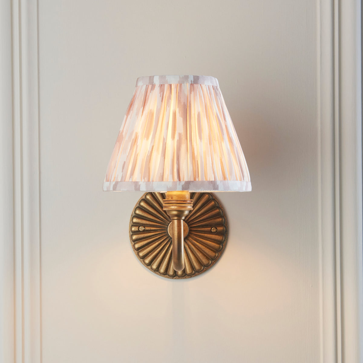 Endon Dahlia Curve wall light lit on a cream panelled wall, neutral ikat shade centred above the fluted brass backplate