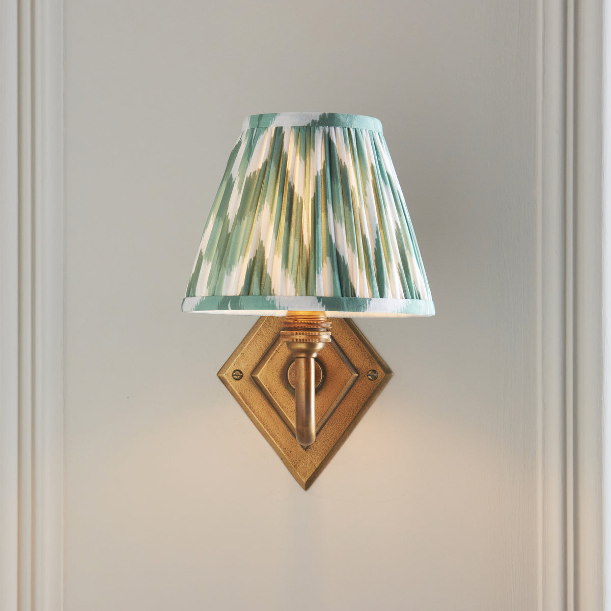 Endon Diamond Curve wall light seen straight on, lit green zigzag pleated shade above the stepped diamond brass backplate on a panelled wall