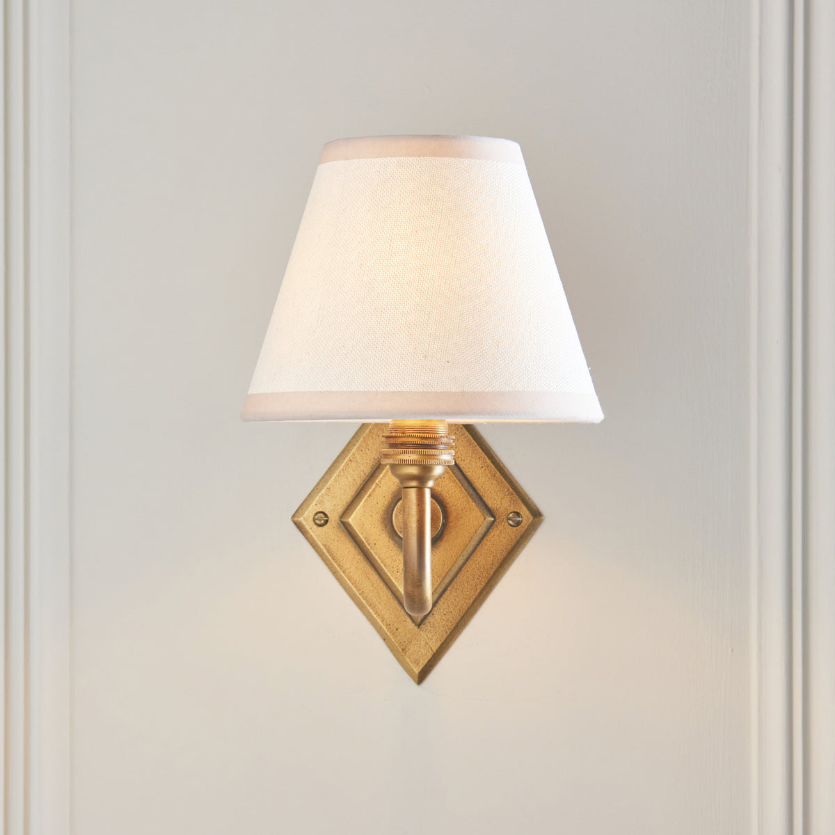 Endon Diamond Curve wall light lit on a cream panelled wall, diamond antique brass backplate below the white fabric shade