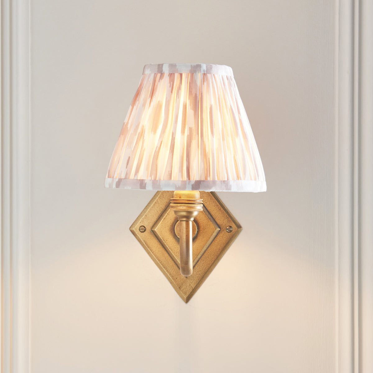 Endon Diamond Curve wall light lit on a cream panelled wall, diamond antique brass backplate sitting directly beneath the pink ikat shade