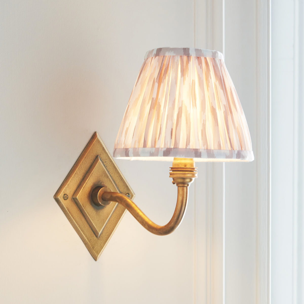 Endon Diamond Curve antique brass wall light lit beside a white window frame, warm glow through the neutral ikat pleated shade