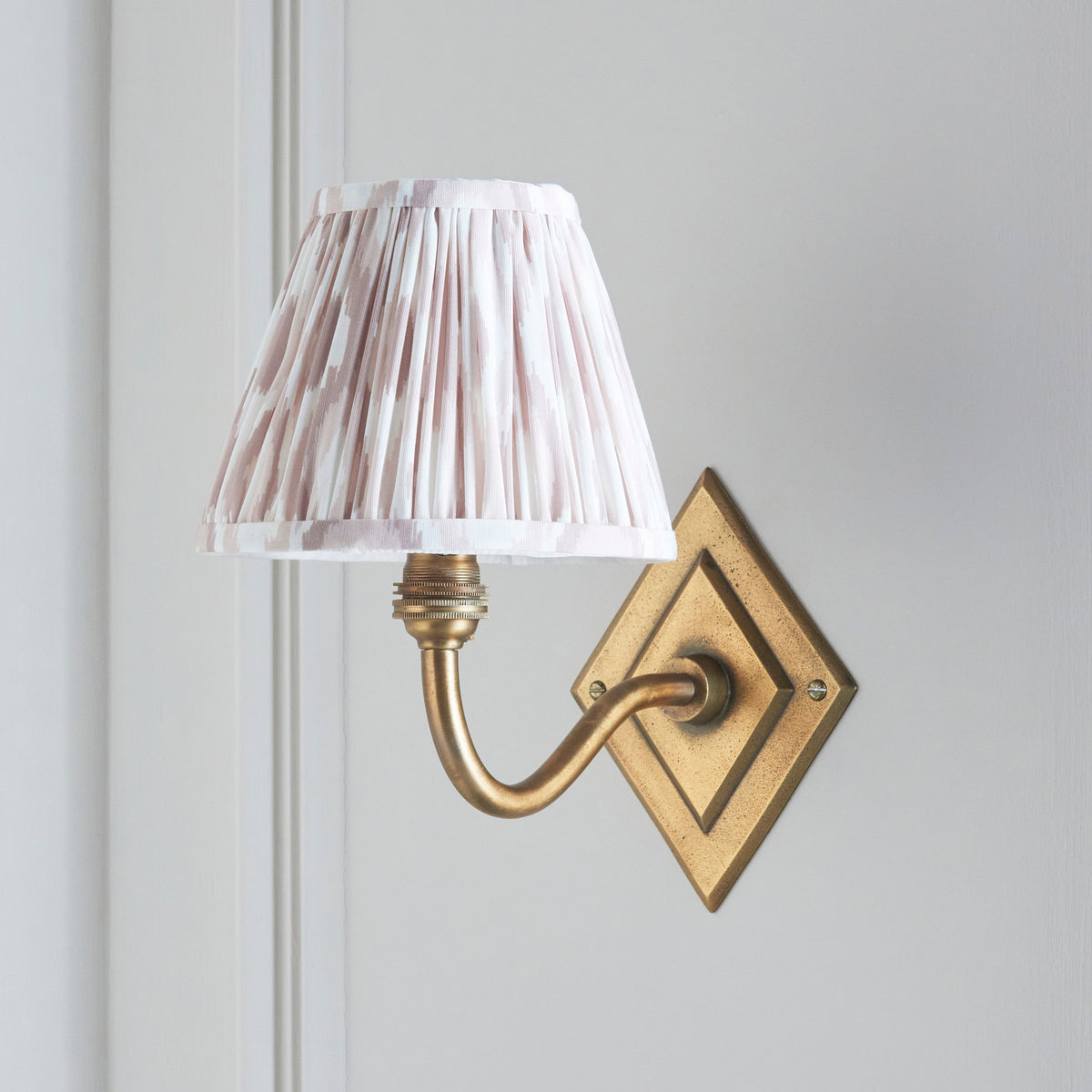 Endon Diamond Curve wall light unlit on a pale grey panelled wall, antique brass arm and blush pink and white ikat shade