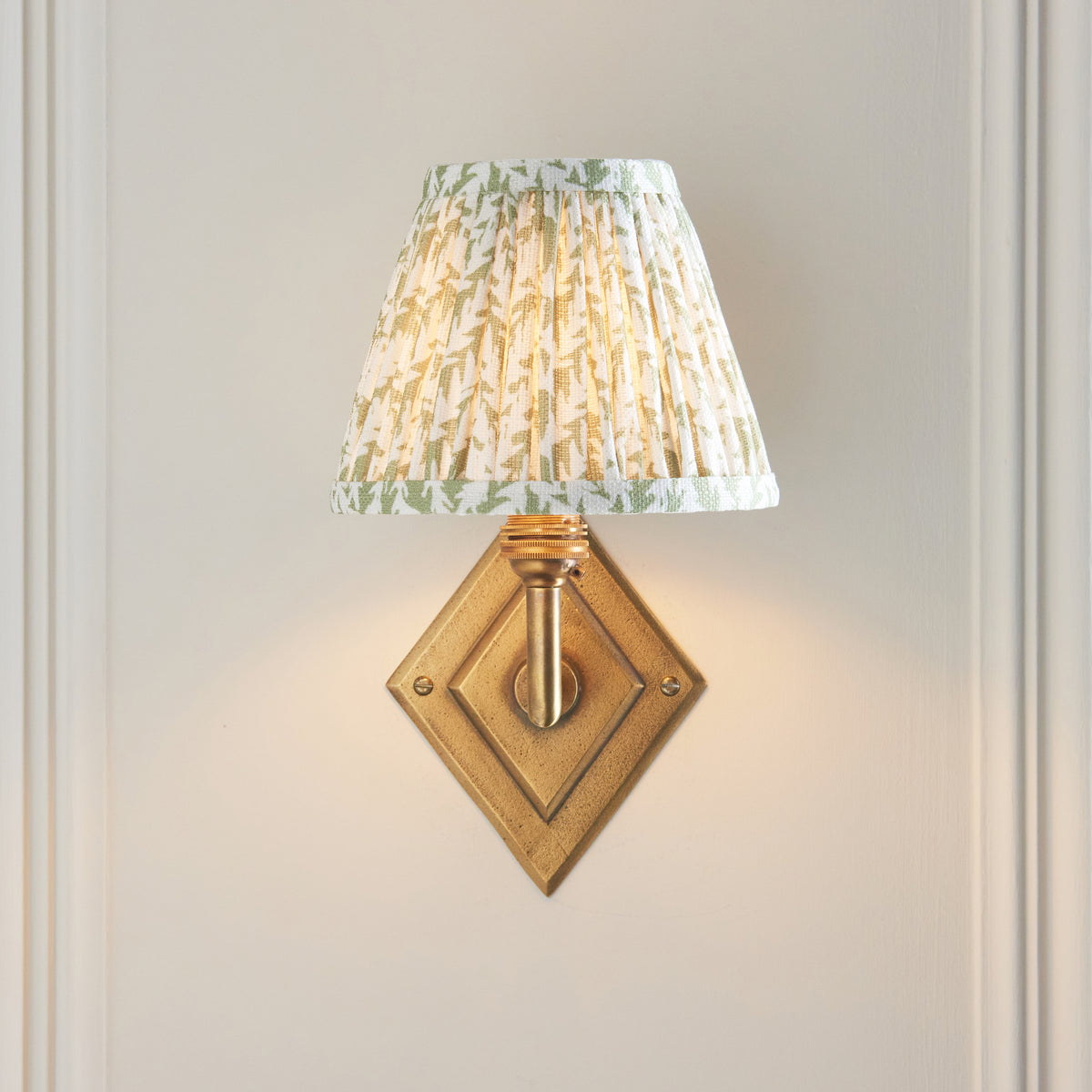 Endon Diamond Angle wall light lit on a cream panelled wall, green leaf pleated shade sitting above the brass diamond backplate