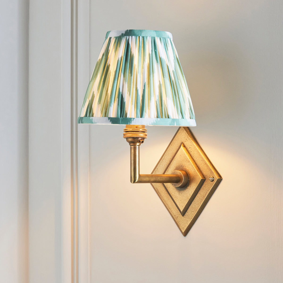 Endon Diamond Angle antique brass wall light lit on a cream panelled wall, green zigzag shade glowing above the diamond backplate