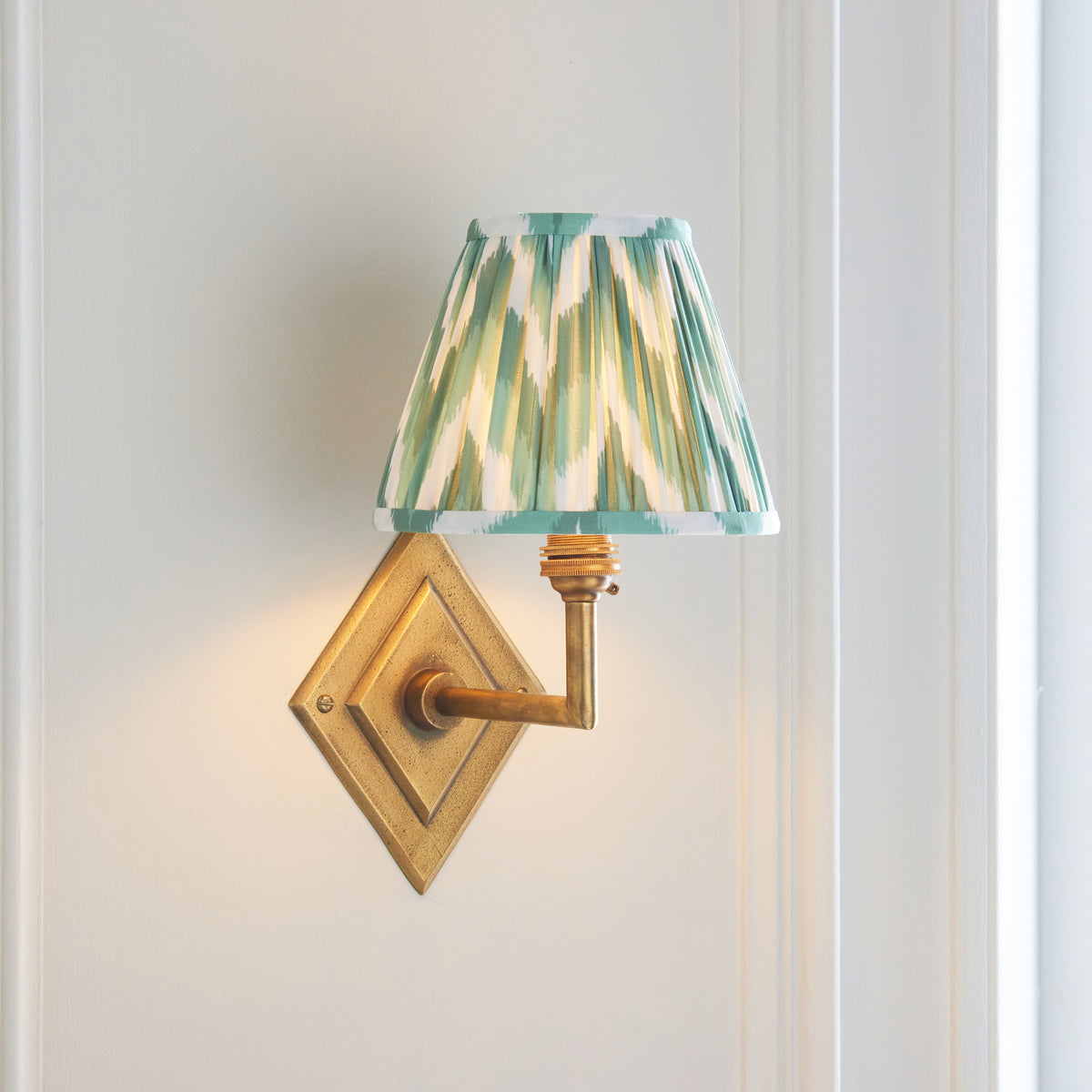 Endon Diamond Angle wall light lit on a cream panelled wall, antique brass arm carrying the Cotswold green zigzag shade out to one side