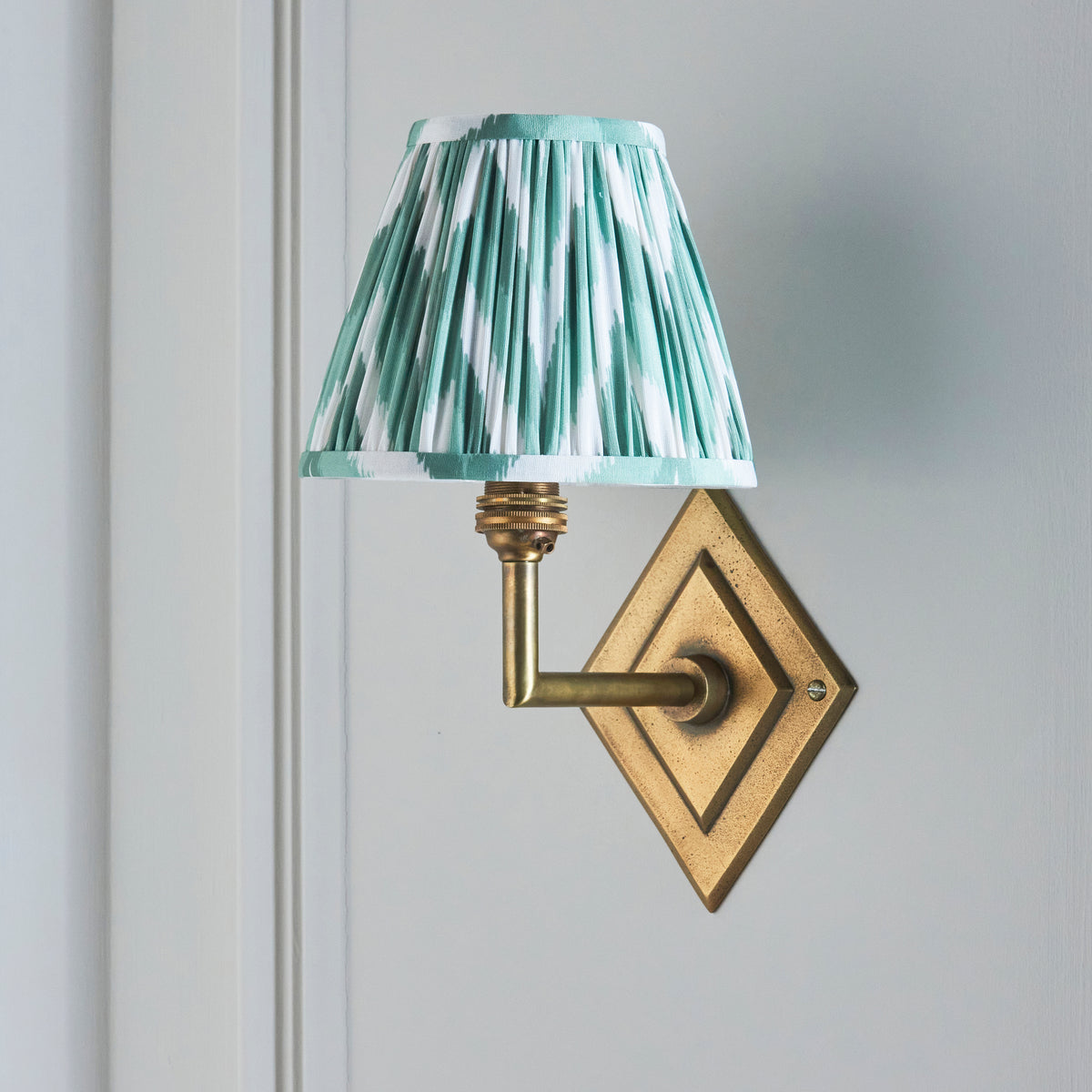 Endon Diamond Angle antique brass wall light unlit on a pale grey panelled wall, showing the green and white zigzag pleated shade