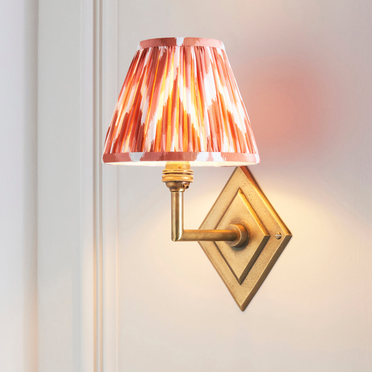 Endon Diamond Angle wall light lit on a cream panelled wall, antique brass diamond backplate with the apricot orange zigzag shade raised to one side