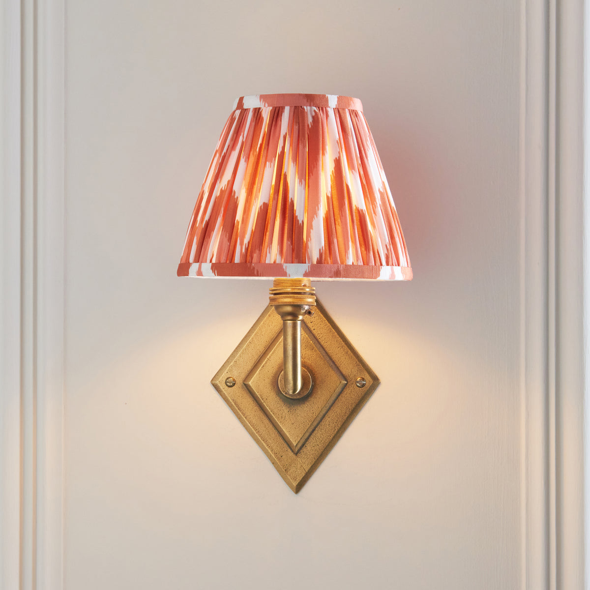Endon Diamond Angle antique brass wall light lit on a cream panelled wall, apricot orange zigzag pleated shade sitting directly above the diamond backplate