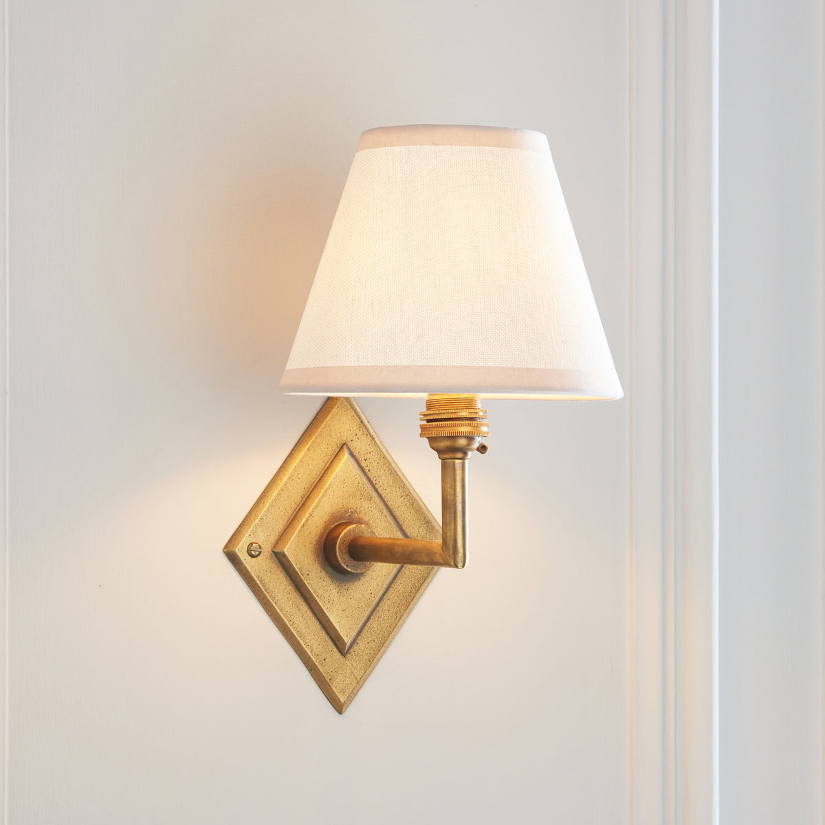 Endon Diamond Angle brass wall light lit, angled arm and vintage white tapered shade casting a warm glow on a pale wall